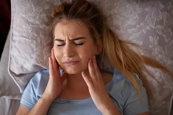 Pained white woman suffering from bruxism and rubbing her jaw while in bed.