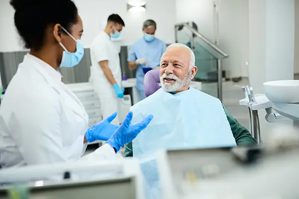 Older white patient sitting in dental chair calmly discussing his oral health with his Black female dentist Durham Prosthodontics dentist visit.